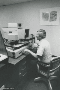 An INS Forensics Document Lab technician examining microfilm reels for evidence of fraud. To his right, on the wall, is an example of a fraudulent document. | Photo credit: USCIS