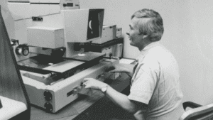 An INS Forensics Document Lab technician examining microfilm reels for evidence of fraud. To his right, on the wall, is an example of a fraudulent document. | Photo credit: USCIS